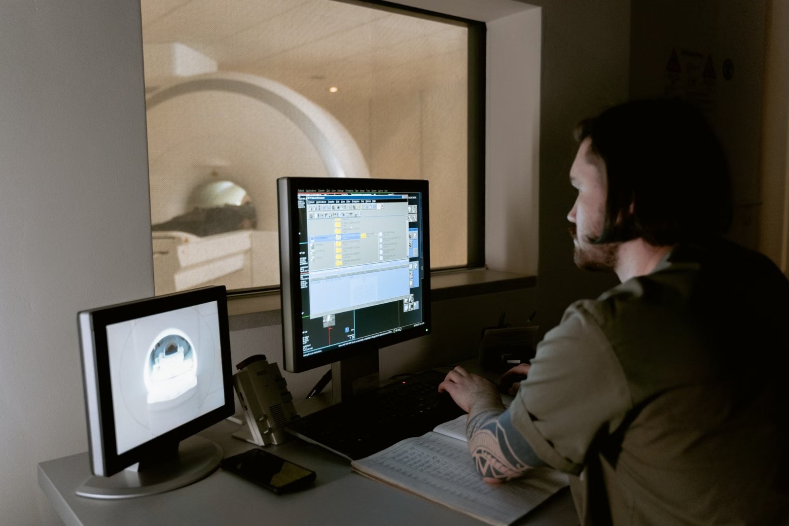 A radiographer at an MRI control console with the scanner behind